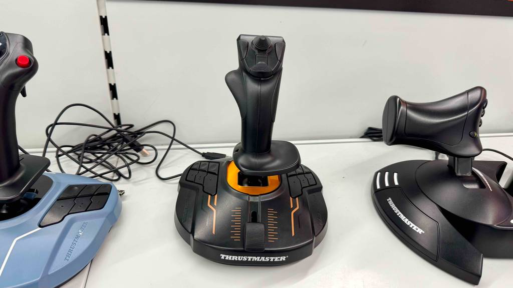 Thrustmaster-Joystick-Test: Das schwarze Modell mit orangefarbenen Verzierungen steht zwischen zwei weiteren Joysticks auf einem weißen Ausstellungsregal.