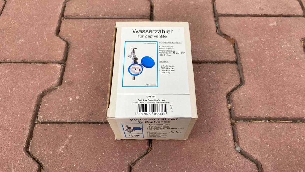 Heller Originalverpackungs-Karton von einem getesteten Wasserzähler für Zapfventile liegt auf einem roten Steinboden.