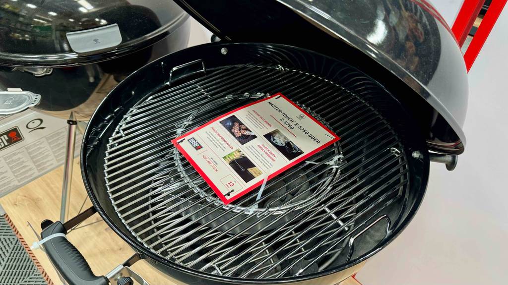 Der runde, getestete Weber-Holzkohlegrill steht mit geöffnetem Deckel und Blick auf das Grillgut-Rost in einem Baumarkt.