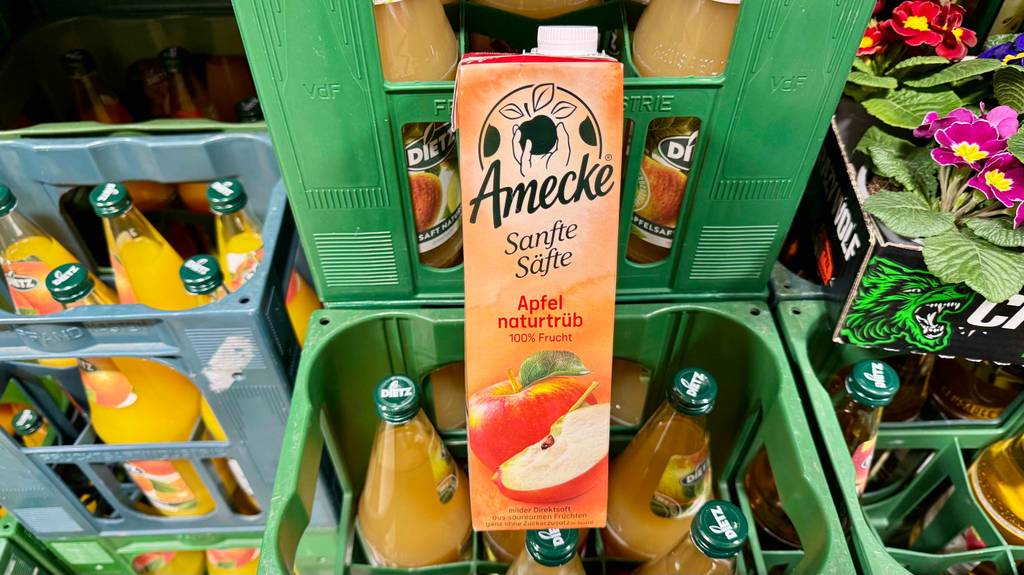 Im Test: Naturtrüber Apfelsaft im Tetrapak der Marke Amecke befindet sich auf einem Kasten mit Fruchtsaftflaschen aus Glas.