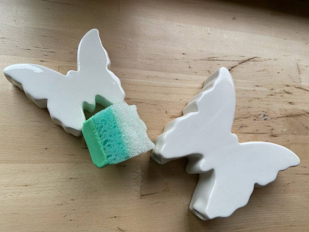 Die grüne, getestete Gesichtsseife inklusive Schwamm liegt zwischen zwei weißen dekorativen Schmetterlingen auf einer Holzoberfläche.