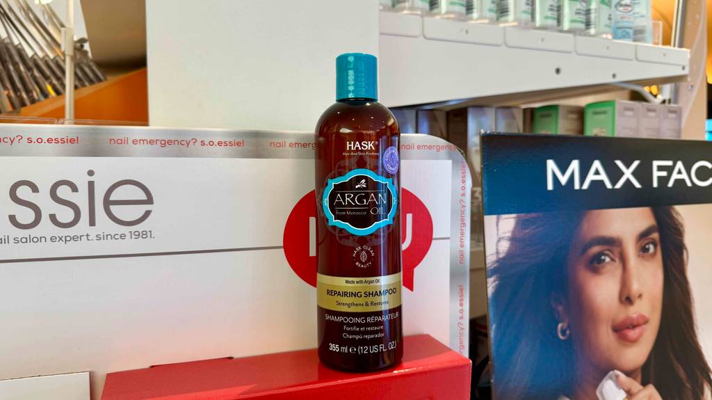 Eine Quetschflasche mit getestetem Hask-Shampoo steht vor Drogerieplakaten auf einer roten Schachtel.
