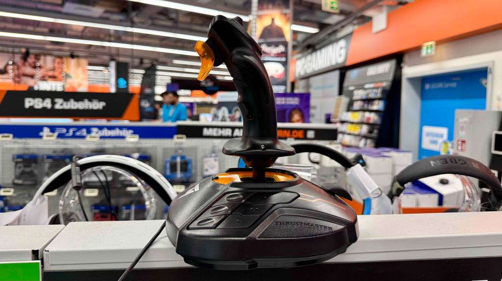Der getestete Thrustmaster-Joystick in Schwarz mit orangefarbenen Highlights steht inmitten eines Elektronikfachmarktes.