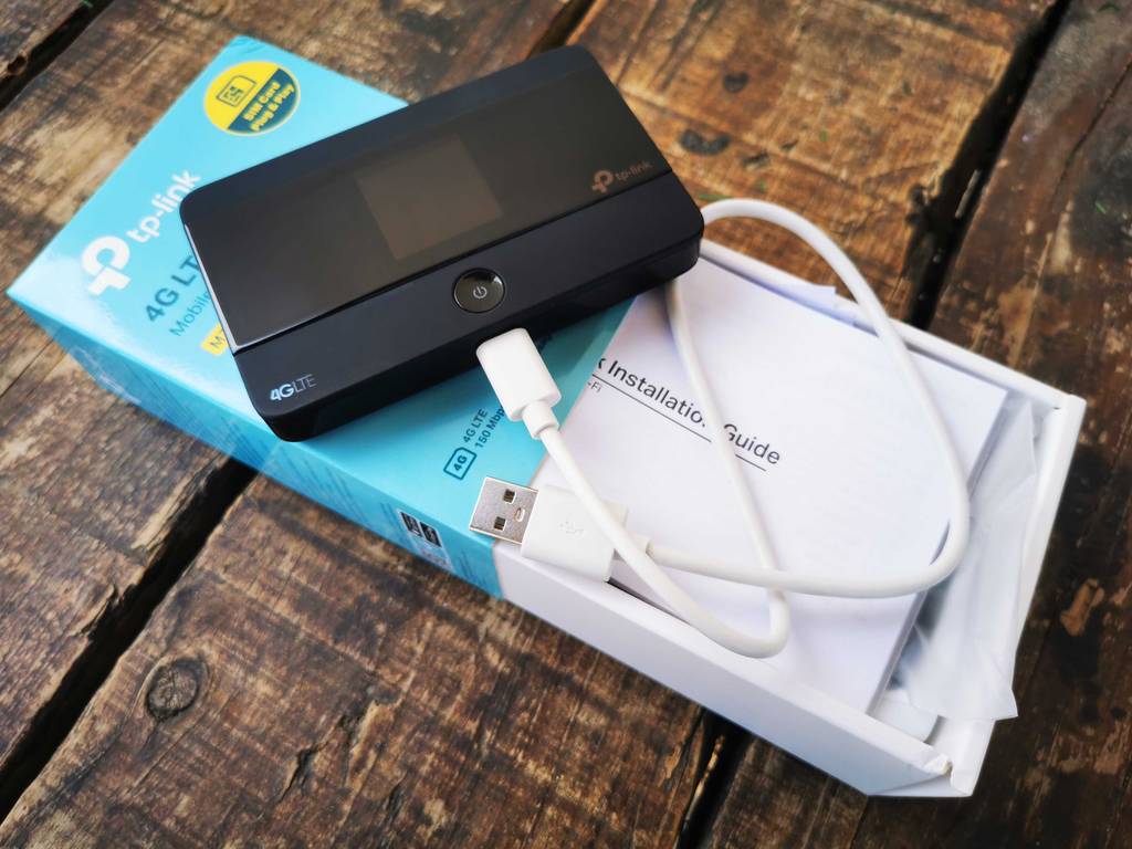 Der schwarze getestete TP-Link-Router liegt inklusive weißem USB-Kabel auf seinem türkisfarbenen, geöffneten Verpackungskarton.