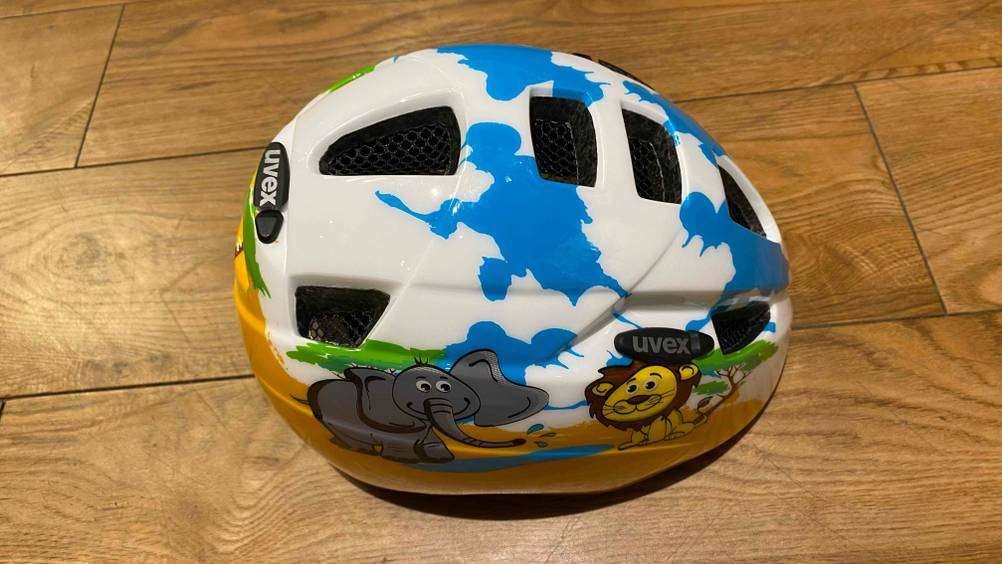 Getesteter Uvex-Fahradhelm für Kinder inklusive niedlichem Tiermotiv, mit Fokus auf die oberen sowie seitlichen Belüftungs-Aussparungen, liegt auf einem Holzboden.