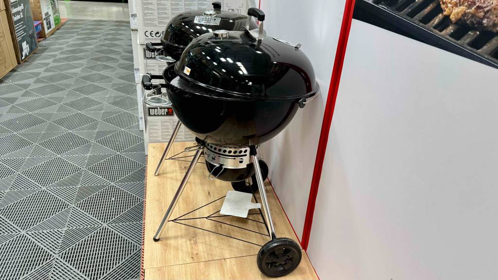Der runde Weber-Holzkohlegrill im Test steht mit geschlossenem Deckel neben einem weiteren runden Standgrill in einem Baumarkt.