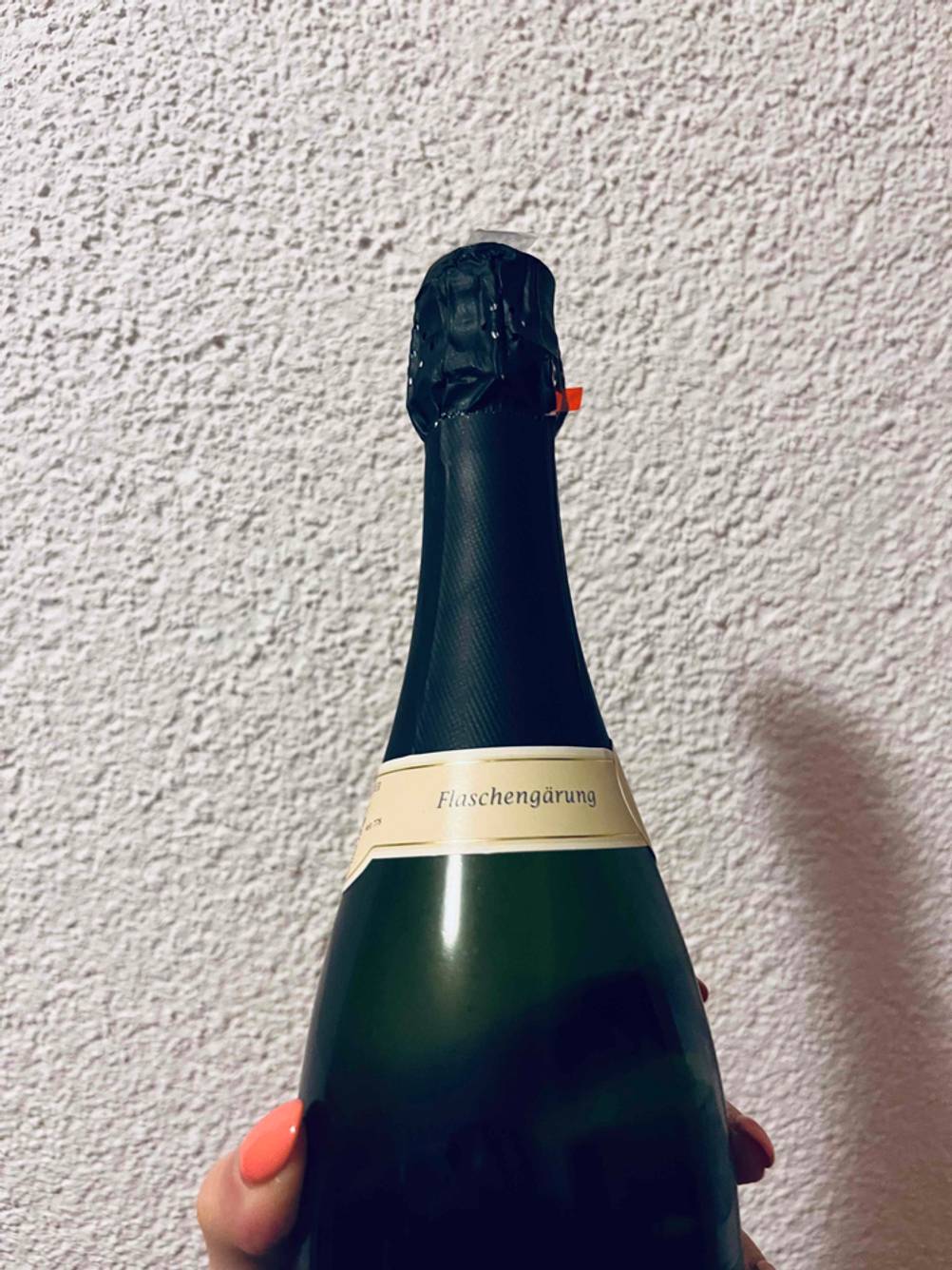 Winzersekt im Test: Hand hält blickdichten Flaschenhals mit heller Flaschengärung-Banderole vor weißer Wand.
