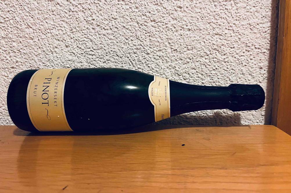 Winzersekt-Test: eine blickdichte Flasche mit Pinot Brut liegt auf Holz vor weißer Wand.