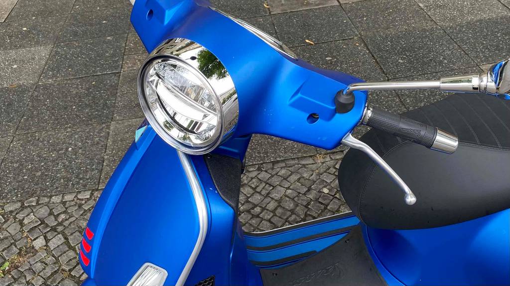 Im Test: Lenker mit rundem Scheinwerfer eines auf der Straße stehenden blauen Vespa Motorrollers.