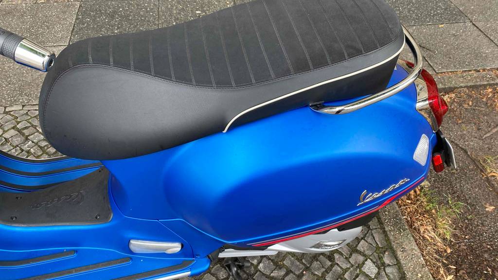 Im Test: schwarzer Sitz eines blauen Vespa Motorrollers, der auf der Straße steht.