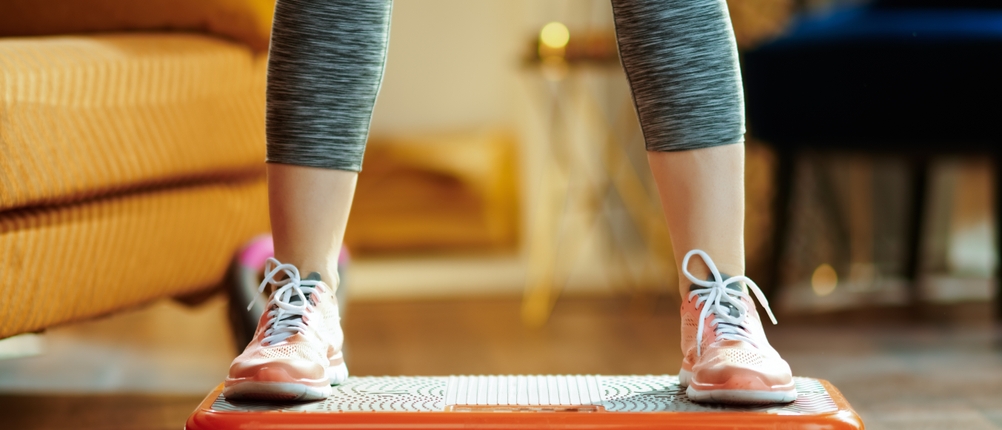 Eine Frau steht mit zwei Beinen auf einer Vibrationsplatte und trägt eine Leggings und Sportschuhe.
