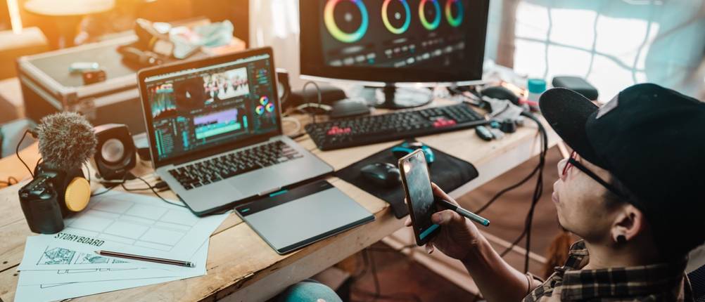 Mann an einem Videoschnitt-Arbeitsplatz mit Laptop, Monitor, Kamera und Smartphone.