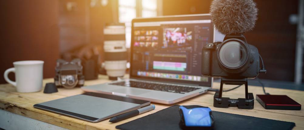 Laptop mit geöffnetem Videoschnittprogramm auf einem Schreibtisch, daneben Kamera mit Mikrofon, Grafiktablett und externe Festplatte.