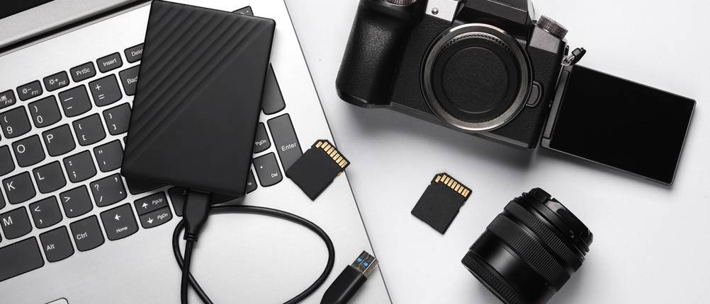 Laptop, externe Festplatte, Kamera, Objektiv und zwei Speicherkarten auf einer hellen Fläche.