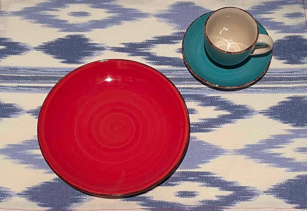 Farbiges getestetes Geschirr bestehend aus einem kleinen roten Teller sowie grünem Unterteller und Tasse, steht auf einer lila-beige gemusterten Tischdecke.