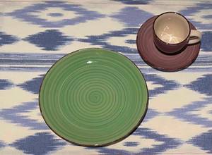 Geschirr-Test: ein kleiner grüner Teller und eine braune Tasse inklusive Unterteller von oben betrachtet auf einer lila-weiß gemusterten Tischdecke.