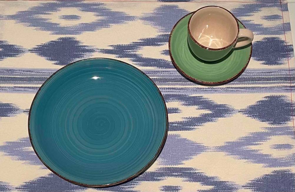 Farbiges getestetes Geschirr bestehend aus einem kleinen grünen Teller sowie Unterteller und Tasse, befindet sich auf einer gemusterten Tischdecke.