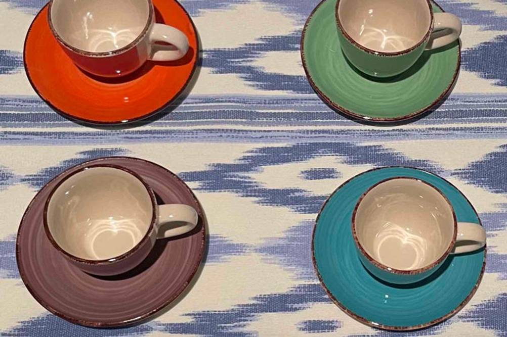 Geschirr im Test: vier Sets mit Tasse und Untertasse in verschiedenen Farben stehen auf blau-weiß gemusterter Fläche.