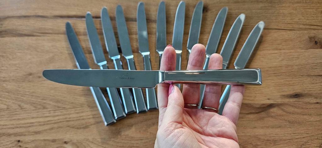 Eine Hand hält ein Messer quer über die anderen als Fächer ausgebreiteten Messer eines getesteten Bestecksets für 12 Personen.