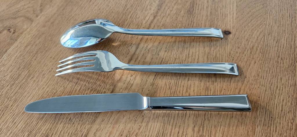 Besteckset für 12 Personen im Test: Je ein Messer, eine Gabel und ein Esslöffel liegen übereinander mit der Rückseite nach oben auf einer Holzfläche.