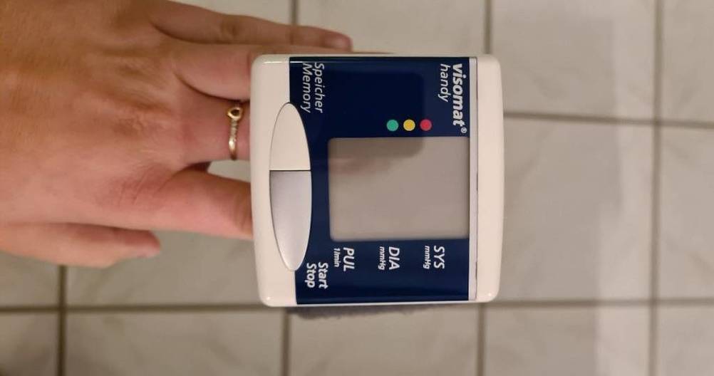 Blau-weißes Visomat Handy Handgelenk-Blutdruckmessgerät im Test wird von einer Hand mit Blick auf die Anzeige vor weißen Fliesen hochgehalten.