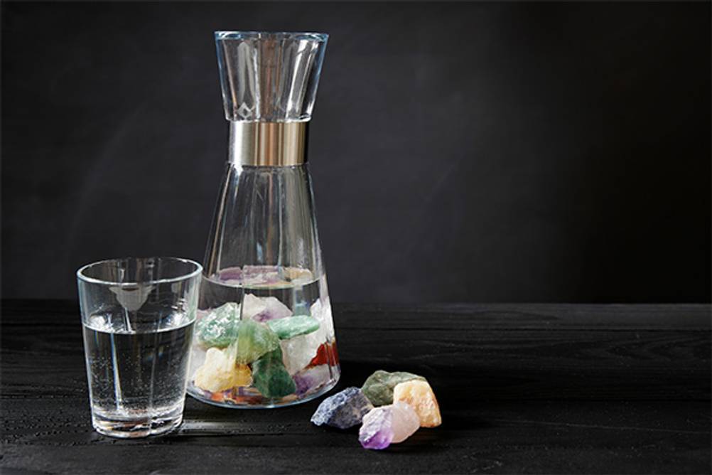 Vitajuwel-Test: Glaskaraffe mit Wasser und Wasserglas stehen vor einem schwarzen Hintergrund. Neben der Glaskaraffe liegen Edelsteine, in der Glaskaraffe liegen einige Edelsteine. 