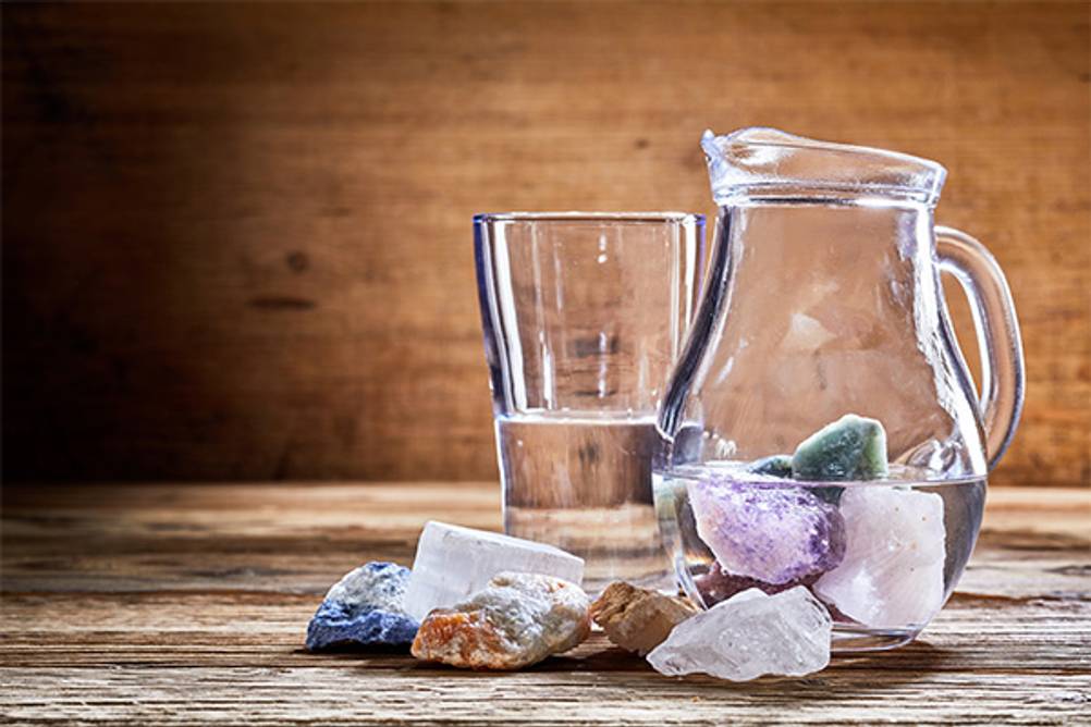 Vitajuwel-Test: Wasserkaraffe und Wasserglas stehen auf einem Holzuntergrund. In der Karaffe befinden sich verschiedene Edelsteine, ebenso vor der Glaskaraffe.