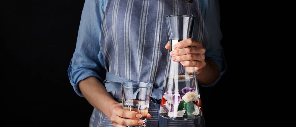 Vitajuwel-Test: Eine Frau mit blauer Bluse und blauer Schürze hält ein Glas mit Wasser sowie eine Glaskaraffe mit Wasser und Edelsteinen in ihren Händen.