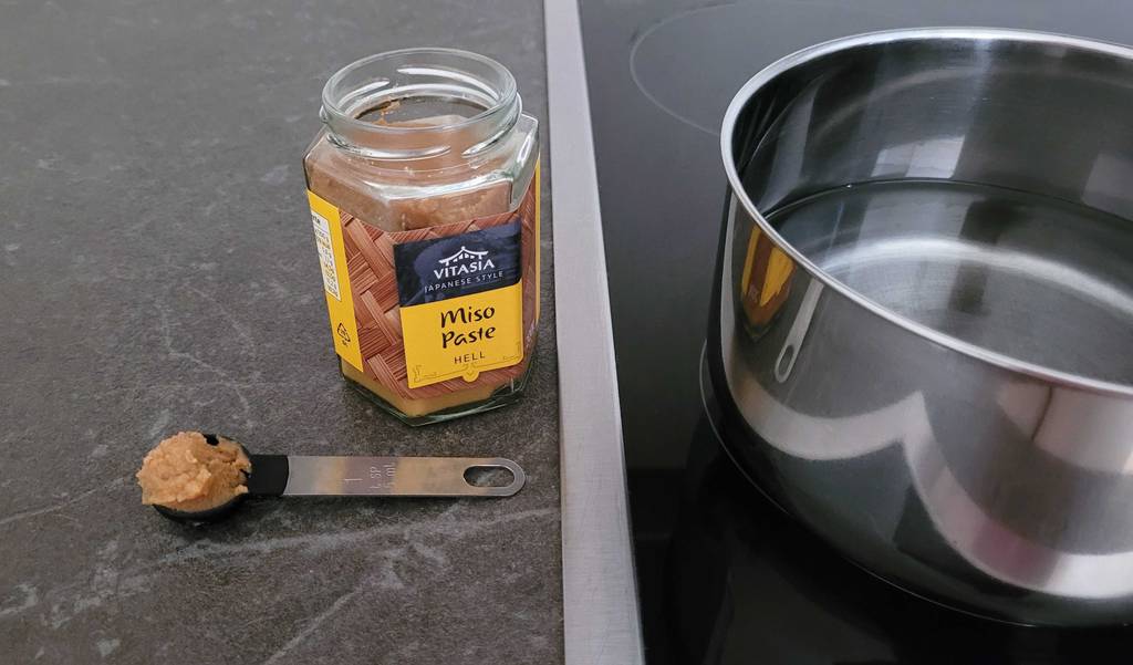 Miso-Paste im Test: Geöffnetes Glas von Vitasia Japanese steht neben einem Topf mit Wasser, davor ein Dosierlöffel voll Paste.