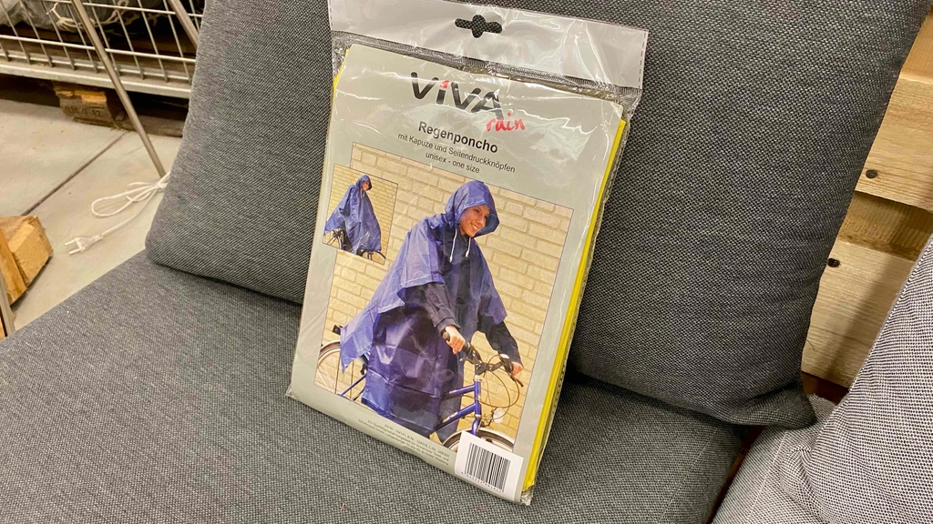 Der lilafarbene Regenponcho im Test befindet sich in der Verpackung auf einem grauen Sofa.