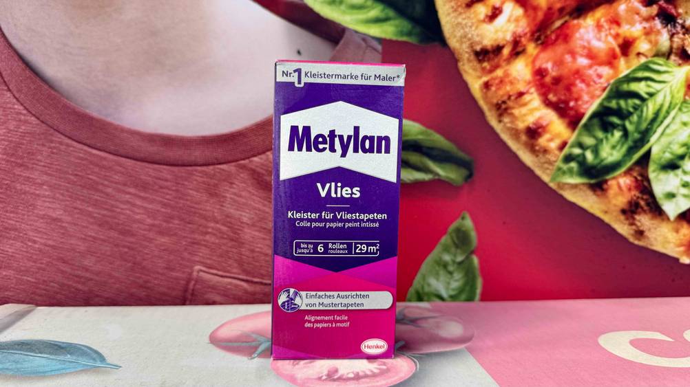 Eine Packung des getesteten Vliestapetenkleisters steht vor einem Plakat mit Pizzadruck.