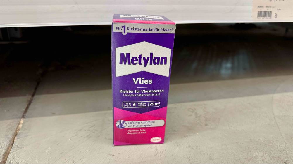 Vor einem Regal steht eine Packung des Vliestapetenkleisters im Test auf dem Boden.