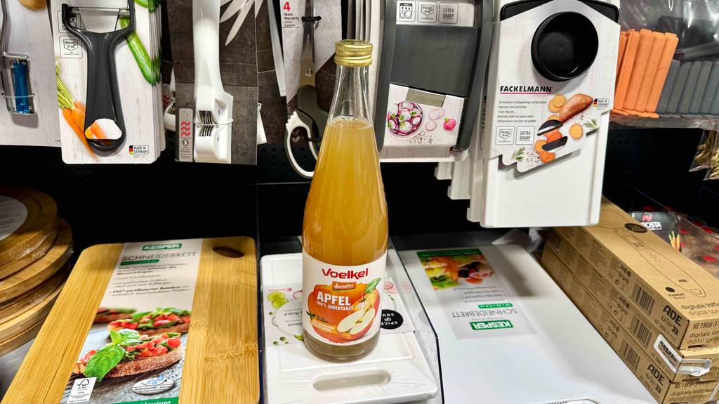 Eine Glasflasche mit Apfelsaft im Test steht auf einem Verkaufsregal vor Küchenutensilien.
