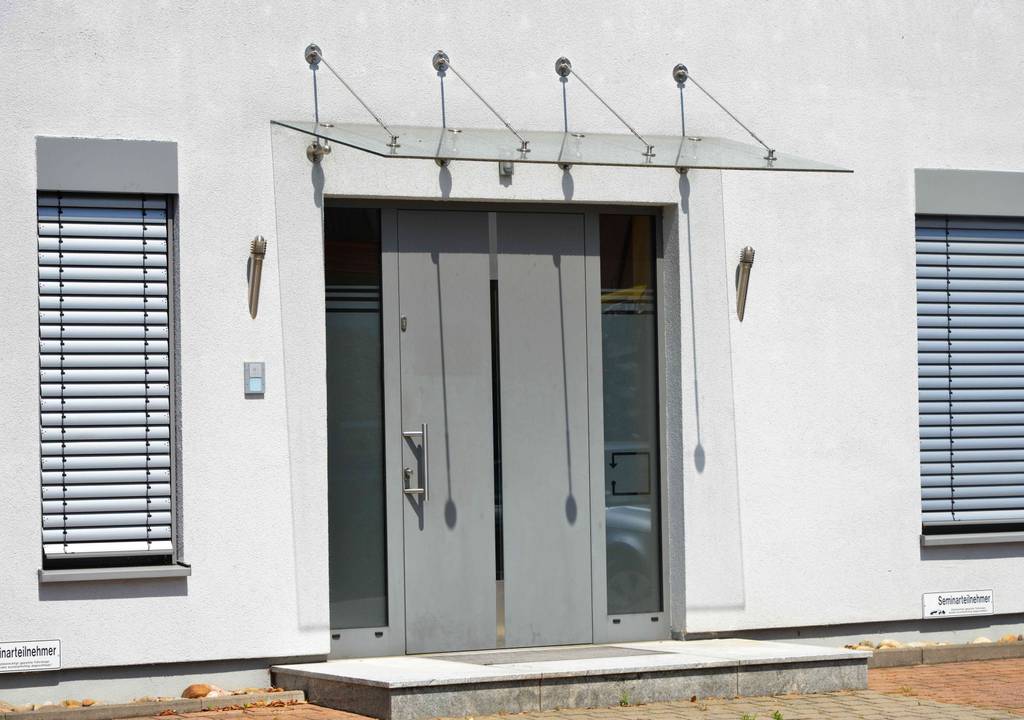 Vordach im Test: Ein Glas-Vordach an einem modernen Haus – gehalten von Edelstahl-Halterungen, die das Dach überwiegend von oben tragen.