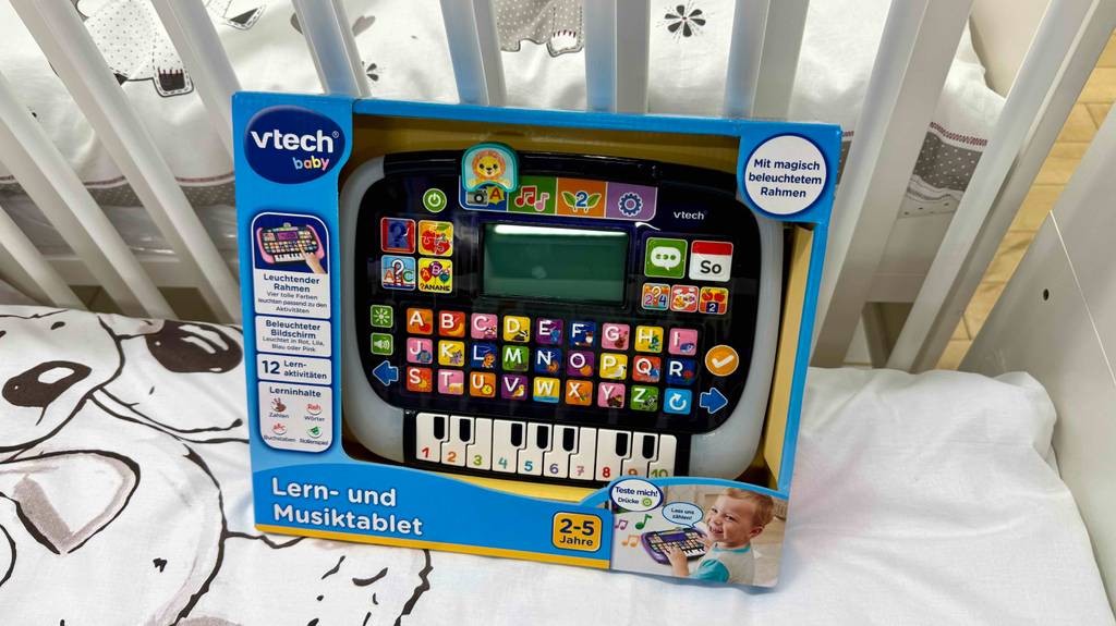 Das Lernspielzeug ab 2 Jahre (im Test) in der blauen Verpackung mit Sichtfenster steht auf einem Kinderbett.