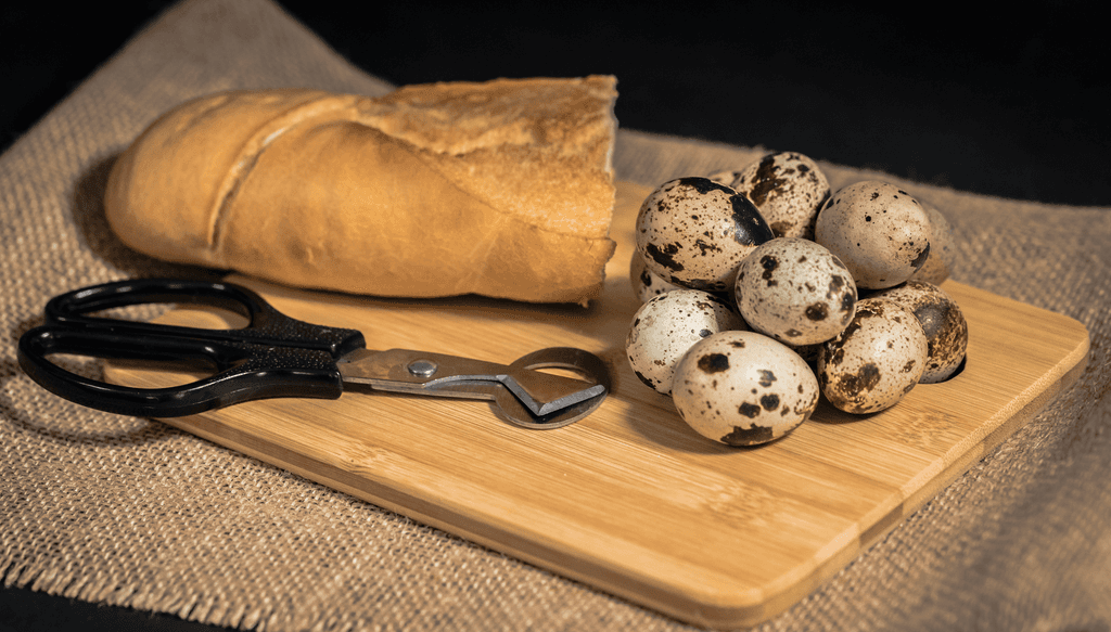 Wachteleierschere-Test: Eine Wachteleierschere, ein Stück Baguette und mehrere Wachteleier liegen auf einem Holzbrett. 