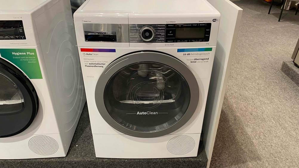 Ein getesteter Wärmepumpentrockner-9-kg von vorne in einer Verkaufsfläche neben anderen Geräten.