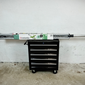 Die verpackte Brabantia Lift-O-Matic 50 liegt quer auf einem schwarzen Rollcontainer im Keller.