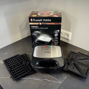 Das getestete Waffeleisen von Russell Hobbs steht zusammen mit der Verpackung und den Wechselplatten auf einer dunklen Arbeitsfläche.