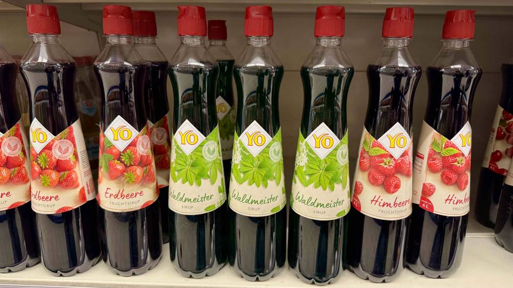 Mehrere Flaschen des Waldmeistersirups im Test in einem Verkaufsregal neben anderen Produkten.
