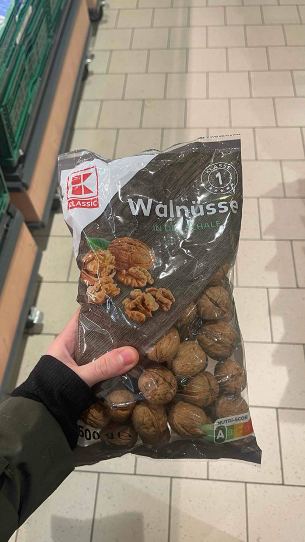 Die Walnüsse mit Schale im Test befinden sich in einer transparenten Tüte der Marke Kaufland, die von einer Hand über einem weißen Fliesenboden gehalten wird.
