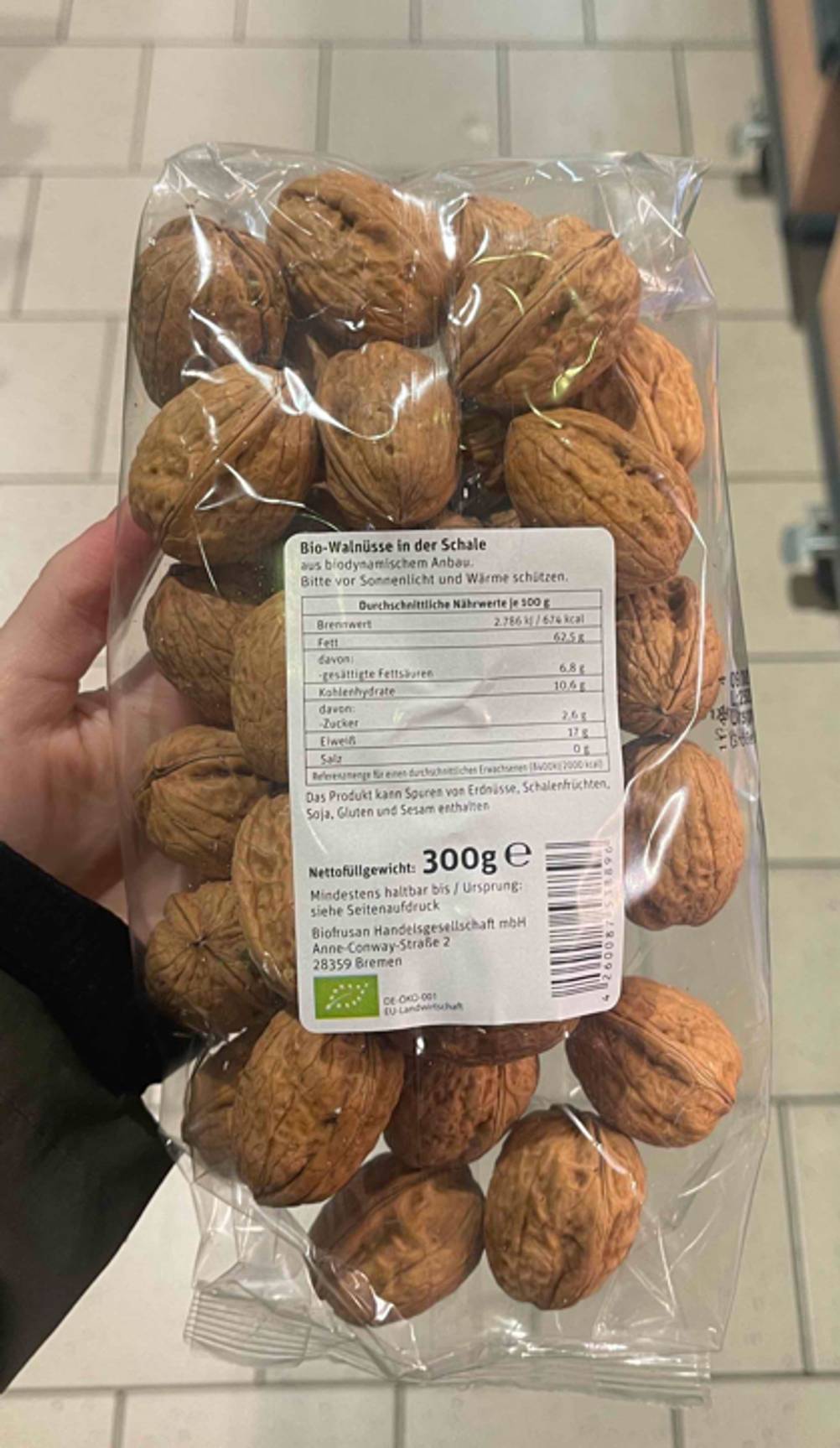 Nahaufnahme der Hand, die die getesteten Walnüsse mit Schale in Bioqualität innerhalb einer transparenten 300-g-Tüte über einem weißen Fliesenboden hält.