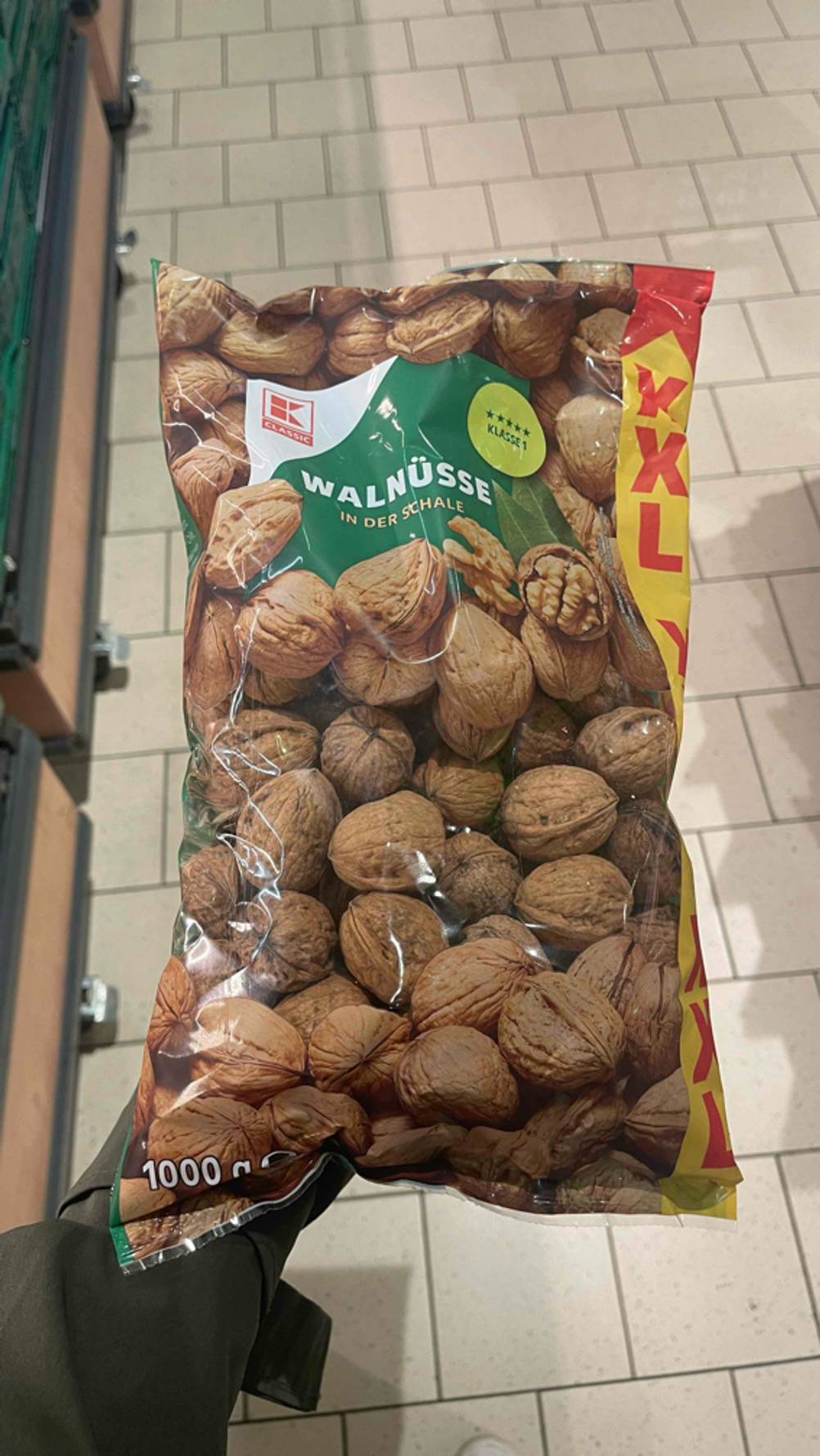 Eine Hand hält die getesteten Walnüsse mit Schale innerhalb einer transparenten 1000-g-Tüte der Marke Kaufland über einem weißen Fliesenboden.
