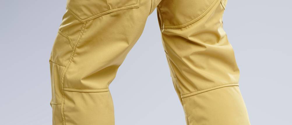 Ein Mann trägt eine gelbe Wanderhose mit markantem Schnitt und aufgesetzten Taschen. Die Hose ist von hinten zu sehen und reicht bis über die Wanderschuhe.