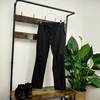 Die Maier Sports Nil Wanderhose hängt an einem Kleiderbügel vor einer Holz-Garderobe. Darunter stehen Wanderschuhe, und eine grüne Pflanze ist daneben platziert.