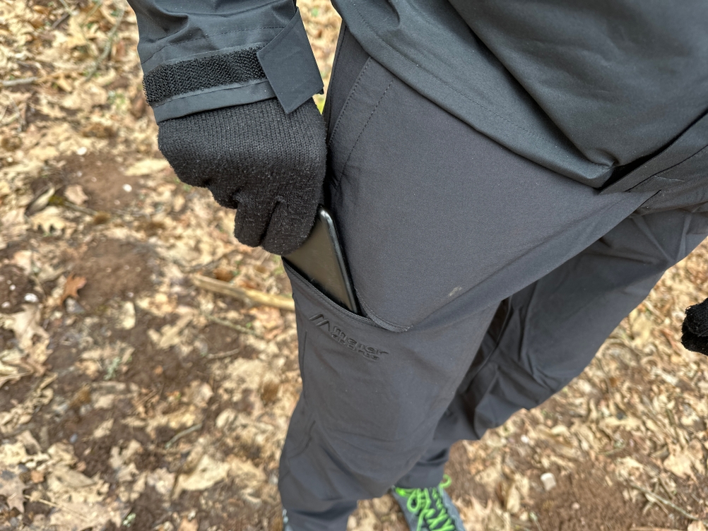 Eine Hand steckt ein Smartphone in die seitliche Tasche der Maier Sports Nil Wanderhose. Die Tasche bietet schnellen Zugriff und sicheren Halt.