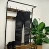 Die RevolutionRace Herren GPX Pro Hose hängt auf einem Kleiderbügel vor einer Holz-Garderobe. Darunter stehen Wanderschuhe, daneben eine grüne Pflanze.