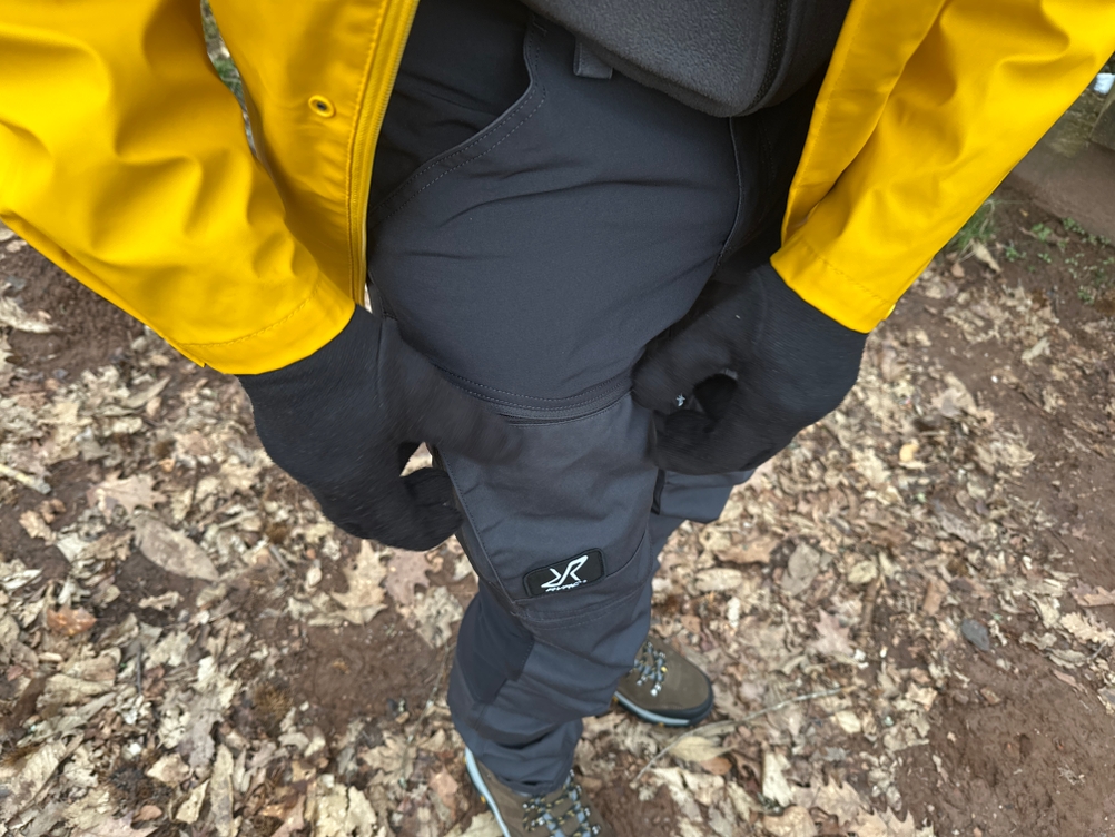 Die RevolutionRace Herren GPX Pro Hose besitzt zwei große Beintaschen mit Reißverschlüssen. Das Logo der Marke sticht hell auf dunkler Hose heraus.