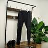 Die Vaude Herren Farley Stretch T-Zip Hose III hängt an einem Kleiderbügel vor einer Holz-Garderobe. Darunter stehen schwarze Wanderschuhe, daneben eine grüne Zimmerpflanze.
