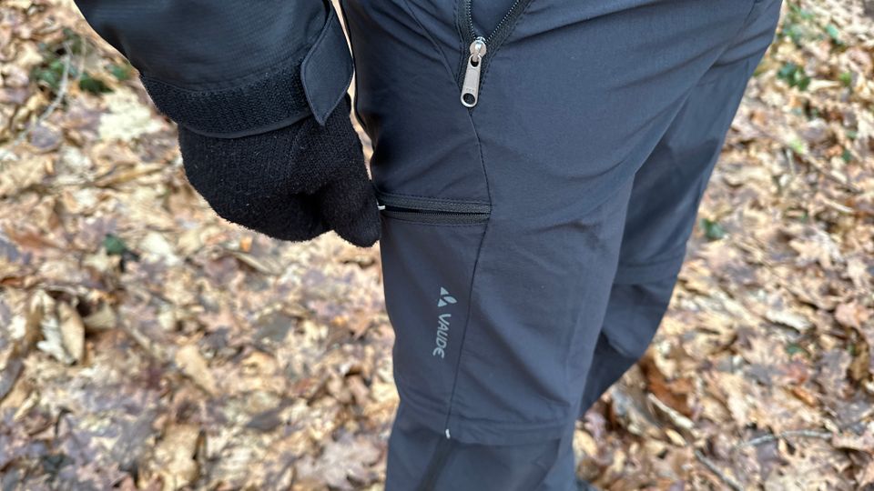 Eine Hand öffnet den seitlichen Reißverschluss der Vaude Herren Farley Stretch T-Zip Hose III. Die verschließbare Tasche sorgt für sicheren Stauraum unterwegs.